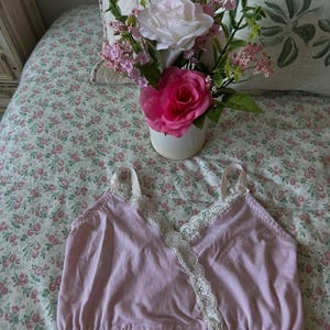 Puede incluir: Un camisón rosa claro con ribete de encaje blanco en el escote y las correas. El camisón está hecho de una tela suave y tiene un dobladillo elástico. Un ramo de rosas rosas y blancas en un jarrón blanco sobre una colcha floral.