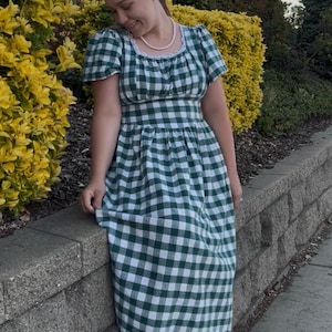 May include: A green and white gingham dress with a square neckline and short sleeves. The dress has a fitted bodice and a flowing skirt. The person is wearing a pearl necklace and white sandals. The dress is made of a lightweight fabric.