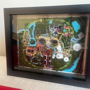 May include: A framed, three-dimensional map of a theme park, featuring detailed miniature buildings, water features, and lush green landscaping. The map is encased in a black frame, creating a shadowbox effect.