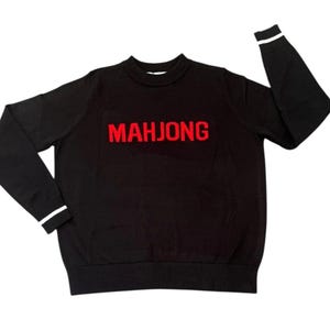May include: Black crew neck sweater with the word "MAHJONG" in red block letters across the chest. The cuffs of the sleeves have a white stripe. The sweater is a casual garment.