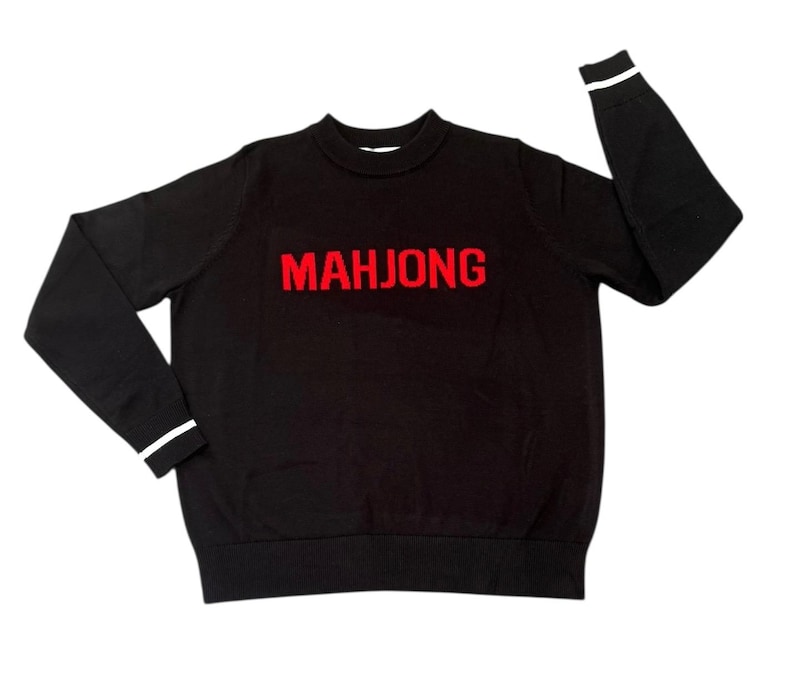 May include: Black crew neck sweater with the word "MAHJONG" in red block letters across the chest. The cuffs of the sleeves have a white stripe. The sweater is a casual garment.