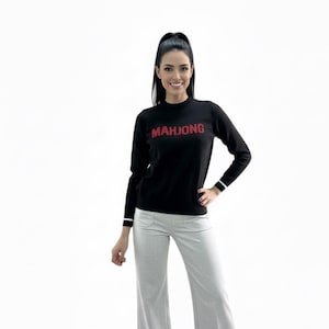 May include: Black long-sleeve sweater with the word "MAHJONG" in red letters. The sweater has white stripes on the cuffs. The model is wearing light gray pants.