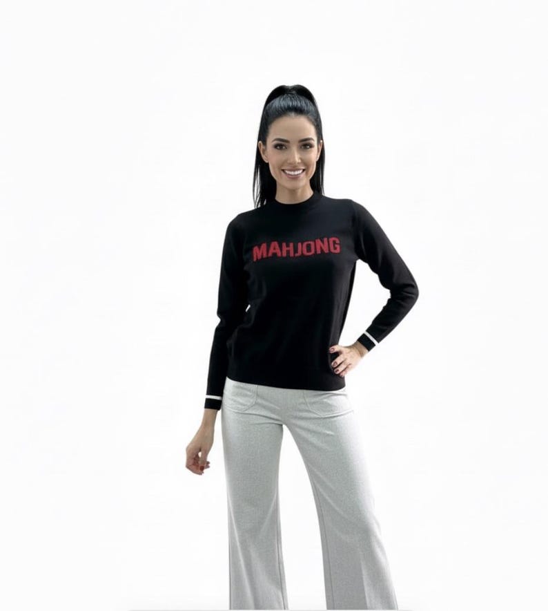 May include: Black long-sleeve sweater with the word "MAHJONG" in red letters. The sweater has white stripes on the cuffs. The model is wearing light gray pants.