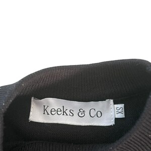 May include: Black knit top with a ribbed turtleneck collar. A white label with the text "Keeks & Co" is sewn into the collar. The label also has a small "XS" size tag.
