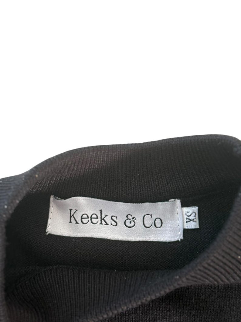 May include: Black knit top with a ribbed turtleneck collar. A white label with the text "Keeks & Co" is sewn into the collar. The label also has a small "XS" size tag.