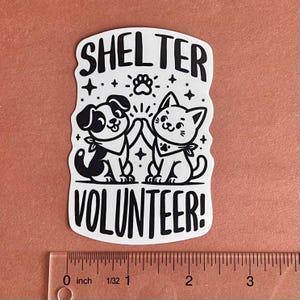 May include: White sticker with black text and illustrations. The text reads "Shelter Volunteer!" with a paw print above the word "Shelter". A black and white cartoon dog and cat are high-fiving each other.