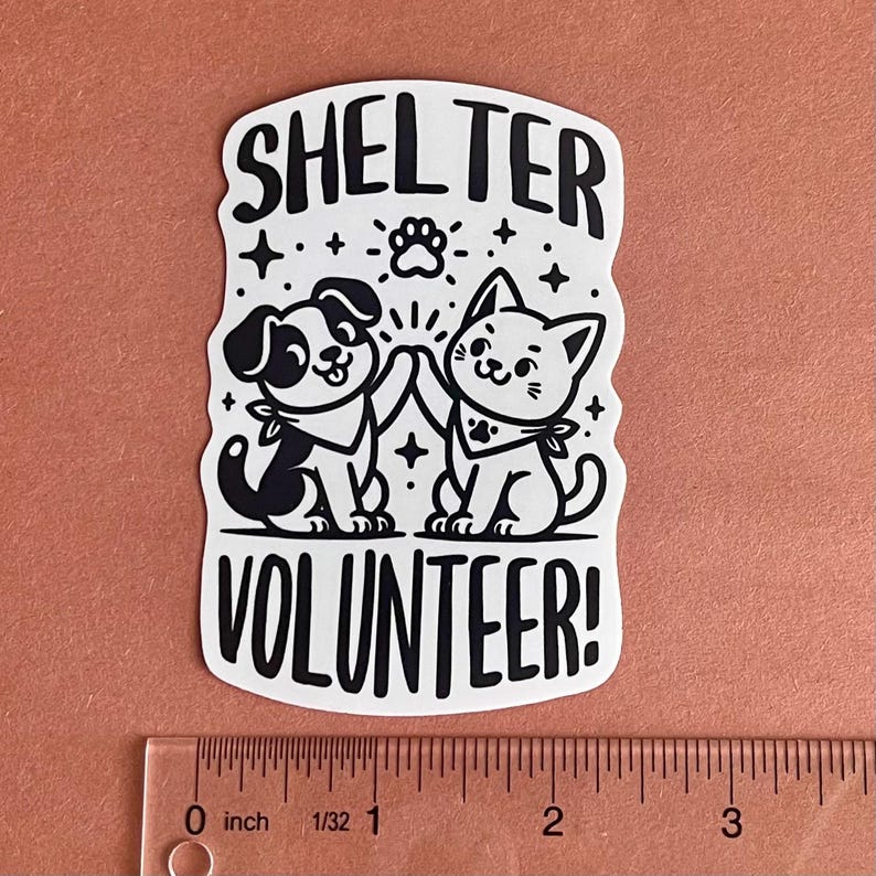 May include: White sticker with black text and illustrations. The text reads "Shelter Volunteer!" with a paw print above the word "Shelter". A black and white cartoon dog and cat are high-fiving each other.