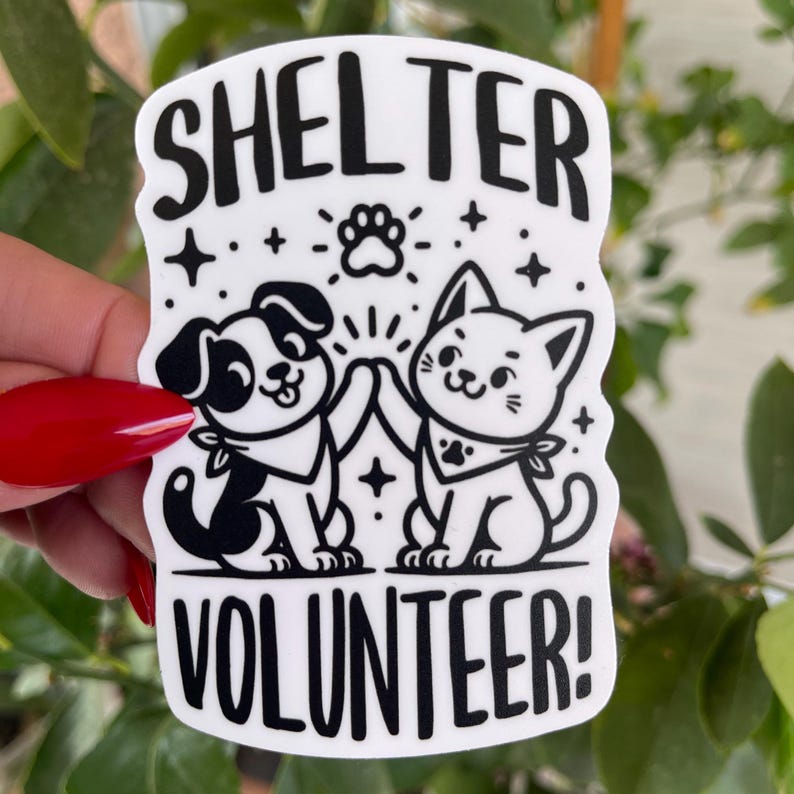 May include: Black and white sticker with a dog and cat high-fiving each other. The sticker says "Shelter Volunteer!" with stars and paw prints around the animals.