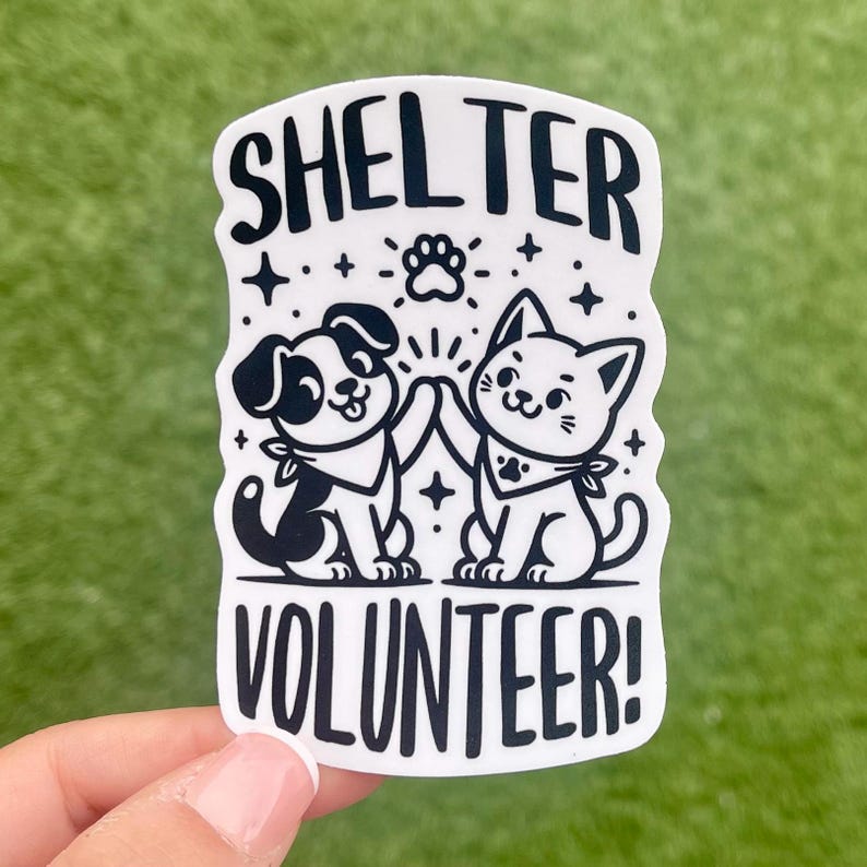 May include: White sticker with black text and illustrations. The text reads "Shelter Volunteer!" with a cartoon dog and cat high-fiving each other.