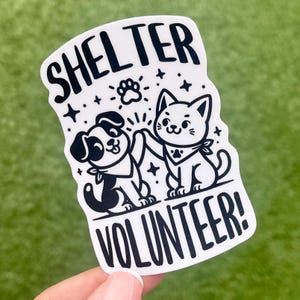May include: White sticker with black text and illustrations. The text reads "Shelter Volunteer!" with a paw print above the word "Shelter". A black and white cartoon dog and cat are high-fiving each other.