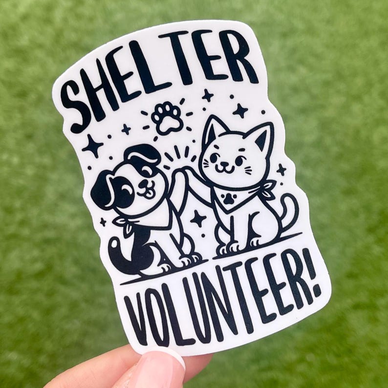 May include: White sticker with black text and illustrations. The text reads "Shelter Volunteer!" with a paw print above the word "Shelter". A black and white cartoon dog and cat are high-fiving each other.