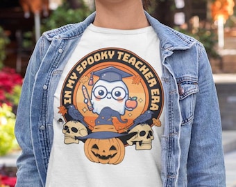 May include: A white t-shirt featuring a graphic of a ghost wearing a graduation cap and holding a pencil and apple. The text surrounding the ghost reads "In My Spooky Teacher Era".