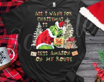 May include: Black t-shirt featuring a Grinch character in a Santa hat and red coat, holding a package. The text reads "All I want for Christmas is less Amazon on my route" in green and white lettering. Christmas-themed.