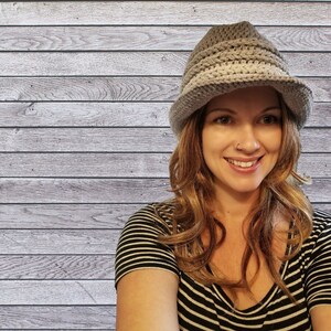 May include: A woman with long brown hair is wearing a gray crocheted hat with a wide brim. She is wearing a black and white striped shirt. The background is a wood panel wall.