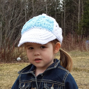May include: A young girl wearing a blue and white crocheted baseball cap, a denim jacket, and a white and blue patterned shirt. She is holding a white butterfly-shaped garden stake.