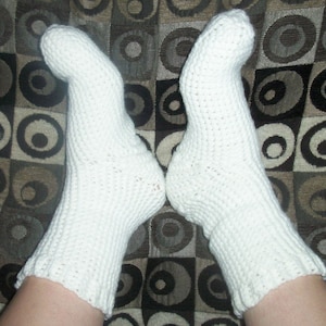 May include: A pair of white crocheted socks with a simple stitch pattern. The socks are shown on a person's feet against a patterned background.