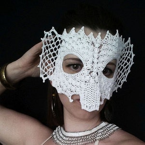 May include: A white, crocheted mask with an intricate, openwork design. The mask covers the eyes and nose, with a jagged edge along the top and sides. The mask is likely handmade and suitable for costume or decorative purposes.