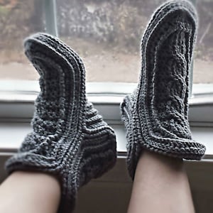 May include: A pair of gray crocheted slipper socks with a cable knit pattern. The socks are worn on a person's feet.