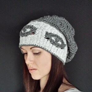 May include: A grey and white crochet beanie hat. The hat features a white band with two grey decorative eye-shaped designs. The top and brim of the hat are grey. The hat is handmade and has a textured appearance.
