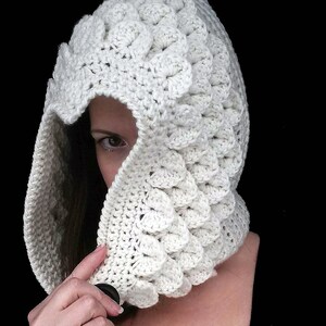 Puede incluir: Cuello con capucha de ganchillo blanco con un patrón texturizado de escamas. El cuello está siendo usado por una persona, con la capucha puesta sobre su cabeza.