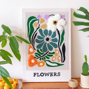 Op de afbeelding: Een textielkunstwerk met een bloemenontwerp. Het heeft een grote witte bloem, een turquoise bloem en oranje accenten. Het woord "FLOWERS" is onderaan geborduurd. Het kunstwerk is omlijst met een neutrale stof.