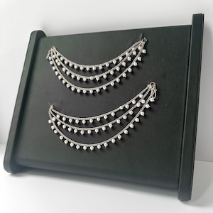 May include: Silver-toned, three-tiered earrings with small, dangling white beads. The earrings are displayed on a black, angled display stand. The earrings have a delicate, elegant design.