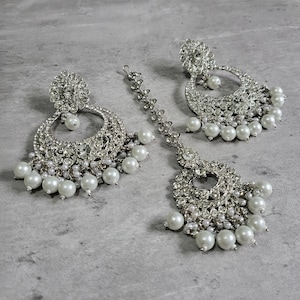 May include: A silver and white bridal jewellery set with pearl accents. The set includes a pair of earrings and a tikka.