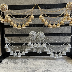 May include: Ornate earrings in gold and silver. The gold earrings have a circular top with pearl detailing and dangling bell charms. The silver earrings have a similar design. Both sets are displayed on a black stand, showcasing the jewellery.