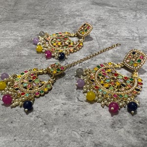 Puede incluir: Un conjunto de pendientes y un adorno para la cabeza dorados y ornamentados con piedras preciosas de colores y perlas de imitación. Las joyas presentan una variedad de colores, incluyendo rojo, amarillo, verde y azul, sobre un fondo gris.