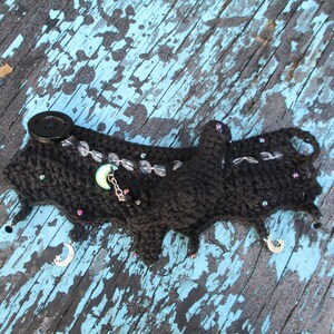 May include: A black crocheted wristband featuring a single black button closure, clear faceted beads, and small dangling silver-toned moon and star charms. The accessory is embellished with tiny iridescent beads scattered across the dark yarn surface.