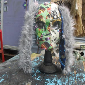 May include: A decorative skull mask with a green and white design featuring cupcakes, bats, and a top hat. The mask has gray fur trim and is adorned with colorful beads. It is mounted on a black stand.