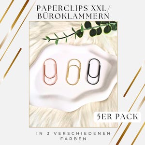 Könnte beinhalten: Drei übergroße Büroklammern in Roségold, Gold und Schwarz, präsentiert auf einer weißen, ovalen Schale. Der Text "PAPERCLIPS XXL / BÜROKLAMMER" steht oben, mit "5ER PACK" und "IN 3 VERSCHIEDENEN FARBEN" unten.