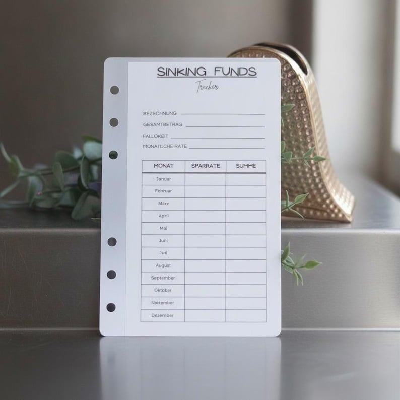 May include: A white paper planner insert with black text titled SINKING FUNDS Tracker. It features fields for BEZEICHNUNG, GESAMTBETRAG, FALLIGKEIT, and MONATLICHE RATE, followed by a table with columns for MONAT, SPARRATE, and SUMME listing months.