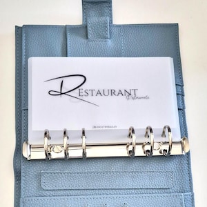Könnte beinhalten: Hellblauer Lederordner mit silbernem Metallringmechanismus. Der Ordner ist geöffnet und zeigt einen weißen Einsatz mit dem Wort "RESTAURANT" in Schreibschrift. Der Einsatz hat auch das Wort "Restaurante" in kleinerer Schrift.