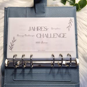 Könnte beinhalten: Ein blauer Lederordner mit silbernen Ringen, der ein transparentes Blatt mit dem Text "JAHRES- CHALLENGE" und "Saving Challenge" enthält. Der Ordner hat einen Klappverschluss und mehrere Kartenfächer.
