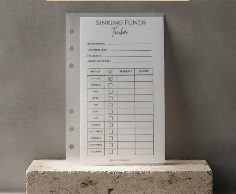 Ein hochkant gestaltetes, transparentes Einlegeblatt für einen Planer mit Ringbindung, beschriftet als „Sinking Funds Tracker“. Das Formular ist auf Deutsch verfasst und dient zum Sparen für bestimmte Ziele. Es enthält Felder für Bezeichnung, Gesamtbetrag, Fälligkeit und monatliche Rate sowie eine Tabelle mit den zwölf Monaten (Januar bis Dezember), Kontrollkästchen, einer Spalte für die Sparrate und einer Spalte für die Summe.