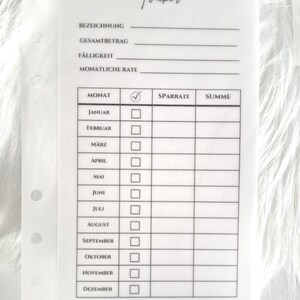 May include: A translucent planner insert titled Sinking Funds Tracker with German text fields for Bezeichnung, Gesamtbetrag, F&auml;lligkeit, and Monatliche Rate. Below, a table lists months from Januar to Dezember with columns for checkmarks, Sparrate, and Summe.