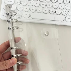 Könnte beinhalten: Transparenter Ordner mit silbernen Metallringen und einem Schnappverschluss. Der Ordner wird vor einer weißen Tastatur gehalten. Der Ordner ist für lose Blätter oder andere Einlagen konzipiert.