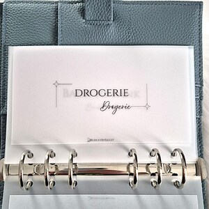 Könnte beinhalten: Ein blauer Lederordner mit silbernen Metallringen, der einen weißen Einsatz mit dem Text "BADROGERIE Drogérie" enthält. Der Ordner ist geöffnet und zeigt transparente Kunststoffhüllen. Der Text "BUDGETBYBAILEY" ist ebenfalls sichtbar.