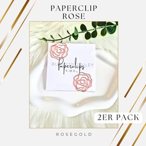 Könnte beinhalten: Roségoldene Büroklammern in Rosenform, präsentiert auf einer weißen Karte mit dem Text "Paperclips" und "Butterly Bailey". Die Karte liegt auf einer weißen Schale mit gewelltem Rand, mit dem Text "2ER PACK" und "Rosegold".