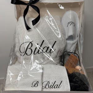 May include: A white gift basket with a clear plastic wrap. The basket contains a white bathrobe, two white towels, a pair of white slippers, a wooden comb, and a black washcloth. The bathrobe, towels, and slippers are embroidered with the name "Bilal".