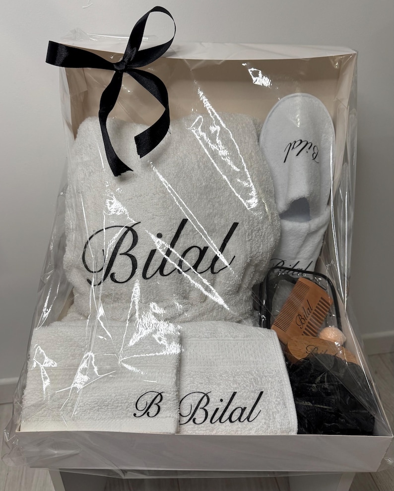May include: A white gift basket with a clear plastic wrap. The basket contains a white bathrobe, two white towels, a pair of white slippers, a wooden comb, and a black washcloth. The bathrobe, towels, and slippers are embroidered with the name "Bilal".