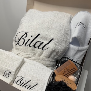 May include: A white bathrobe, two white towels, a pair of white slippers, a black bath pouf, a wooden comb, and a wooden bath brush, all personalized with the name "Bilal".