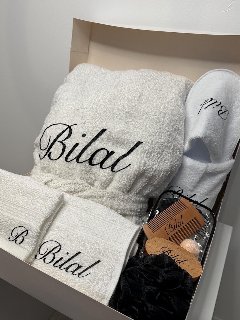 May include: A white bathrobe, two white towels, a pair of white slippers, a black bath pouf, a wooden comb, and a wooden bath brush, all personalized with the name "Bilal".