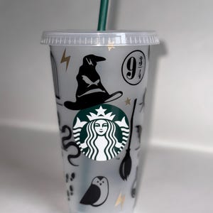 May include: A clear plastic reusable cup with a black and green Starbucks logo and various Harry Potter themed stickers, including a black witch's hat, a snake, a broom, a golden lightning bolt, and the number "9 3/4".