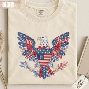 May include: An ivory t-shirt featuring a graphic of a bald eagle composed of various red, white, and blue patterned fabric patches. The eagle holds arrows and an olive branch, with a central shield displaying the American flag. The label reads Comfort Colors.