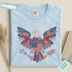 May include: A light blue t-shirt featuring a central graphic of a bald eagle composed of various red, white, and blue patterned fabric patches. The eagle holds arrows and a branch, with the text Chambray and Luvais Designs visible.