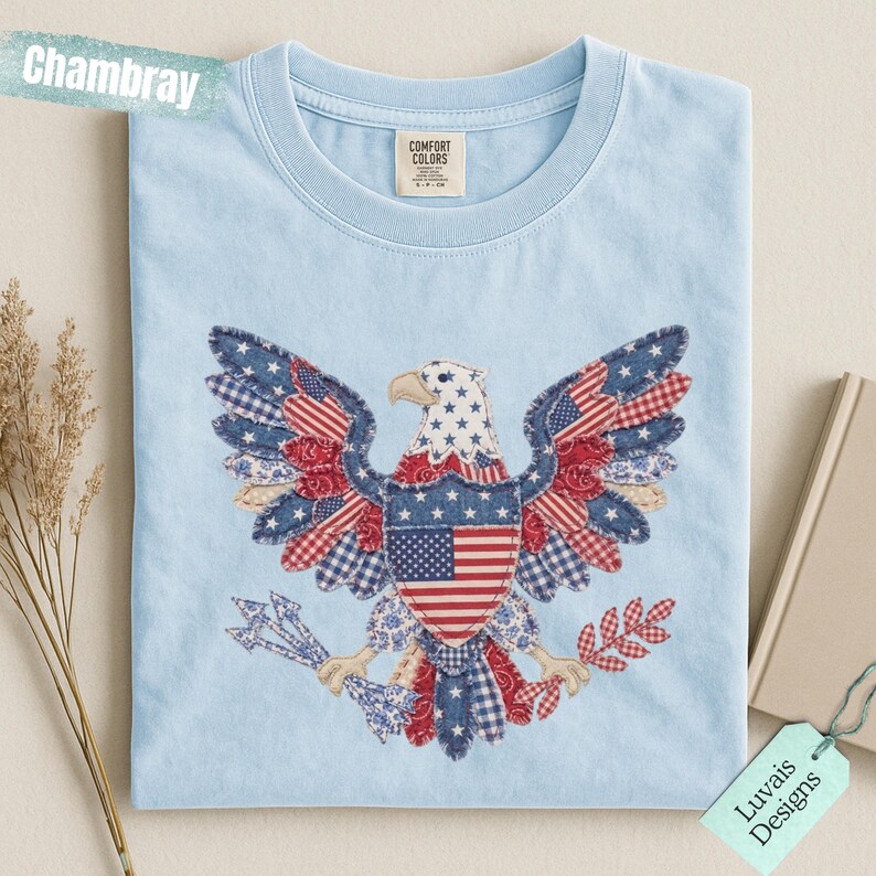May include: A light blue t-shirt featuring a central graphic of a bald eagle composed of various red, white, and blue patterned fabric patches. The eagle holds arrows and a branch, with the text Chambray and Luvais Designs visible.