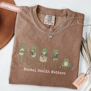 Op de afbeelding: Een espresso-kleurig t-shirt met een afbeelding van vijf groene kikkers die verschillende activiteiten uitvoeren, met de tekst "Mental Health Matters" eronder gedrukt. Het shirt heeft een ronde hals en een Comfort Colors-label in de kraag.