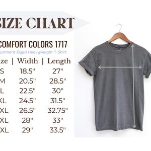 May include: A charcoal gray short-sleeve t-shirt hanging on a wooden hanger, accompanied by a size chart for Comfort Colors 1717 garment-dyed heavyweight t-shirts. The chart lists widths and lengths in inches for sizes S through 4XL.